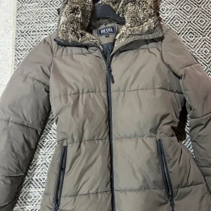 Brun dunjacka med fuskpäls från Jofama - Snygg brun dunjacka från Jofama med mysig fuskpäls i luvan. Jackan har dragkedja framtill och två praktiska fickor med dragkedja. Perfekt för kalla dagar med sin quiltade design och varma känsla.