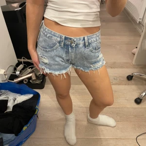 Ljusblå denimshorts med fransig kant - Säljer ett par ljusblå denimshorts med fransig kant och slitna detaljer framtill. Klassisk femficksmodell med låg midja och knappgylf. Perfekta för varma dagar och ger en avslappnad vibe.