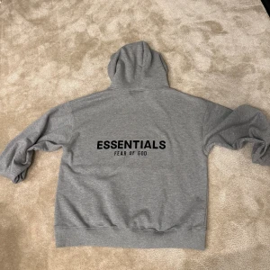 Grå Essentials Fear of God hoodie - Säljer en grå hoodie från Fear of God Essentials med svart logga på ryggen och bröstet. Klassisk känguruficka framtill, ribbade muddar och huva. Perfekt för dig som gillar streetwear och enkel stil.