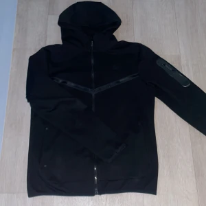 Svart Nike vindjacka med huva M - Svart vindjacka från Nike i storlek M med huva och hel dragkedja framtill. Jackan har Dri-Fit-material som andas och reflekterande detaljer över bröstet. Snygg sportig look med diskret Nike-logga på vänster sida.