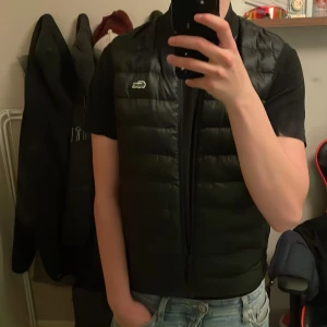 Svart dunväst från Lacoste - Svart dunväst från Lacoste med klassisk krokodillogga på bröstet. Västen har quiltad design, dragkedja framtill och en clean, sportig look. Skick 7/10 då den har en liten skada (se bild 2) men ingenting som syns vid användning, men därav pris. Skriv vid minsta fundering