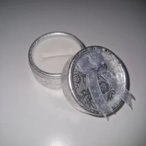 Smyckes ask - Snygg rund ask i silverfärg med blommigt mönster och en transparent rosett på locket. Perfekt för att ge bort en ring/örhängen eller förvara ditt favoritsmycke. Asken har en lyxig känsla och passar dig som gillar stilrena detaljer.