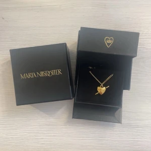 Maria Nilsdotter liknande halsband  - Snyggt halsband från Maria Nilsdotter i guld med ett hjärtformat hänge och pil-detalj. Kedjan är klassisk och passar perfekt till dig som gillar unika smycken med cool design. Levereras i original ask.