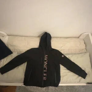 Svart vindjacka från Moncler - Svart vindjacka från Moncler med huva och dragkedja framtill. Stor logotyp i vitt längs dragkedjan och klassisk Moncler-patch på ärmen. Jackan har långa ärmar och två fickor med dragkedja. Perfekt för dig som vill ha en stilren och sportig look.