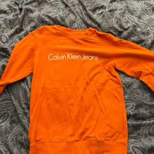 Orange sweatshirt från Calvin Klein Jeans - Säljer en snygg orange sweatshirt från Calvin Klein Jeans med vit logga framtill. Tröjan har rund halsringning, långa ärmar och ribbade muddar vid ärmslut och nederkant. Perfekt för dig som vill sticka ut med färg och stil.