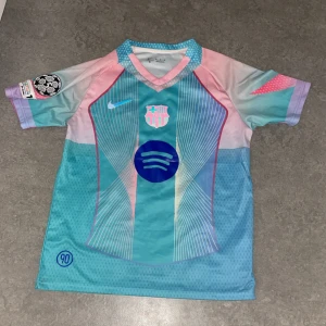 FC Barcelona Lamine Yamal 10 Nike tröja - Säljer en FC Barcelona fotbollströja med Lamine Yamal 10 på ryggen. Tröjan har coola pastellfärger i blått, rosa och lila, med klubbmärke och Champions League-märke på ärmen. Tillverkad i lätt och ventilerande polyester, perfekt för match eller träning.