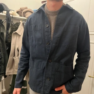 Mörkblå overshirt med knappar - Snygg mörkblå overshirt med fyra stora fickor framtill och klassisk krage. Jackan har raka linjer, knäppning med stora knappar och är tillverkad i bomull. Perfekt för lager-på-lager och enkel att styla till olika outfits. 