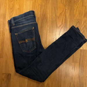 Mörkblå Nudie Jeans - Säljer ett par mörkblå jeans från Nudie Jeans med klassisk femficksdesign och snygga kontrastsömmar på bakfickorna. Jeansen har normal passform och raka ben, tillverkade i slitstark denim av bomull. Perfekta för dig som gillar stilrena och tidlösa jeans. Ny pris 1399