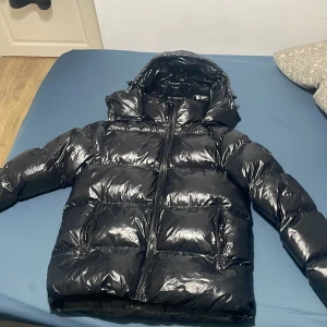Svart pufferjacka från Everest - Säljer en svart, glansig pufferjacka från Everest med huva och dragkedja framtill. Jackan har två stora fickor med dragkedja och en riktigt fet vinterlook. Perfekt för dig som vill hålla stilen när det är kallt ute. Priset kan diskuteras är helt ny