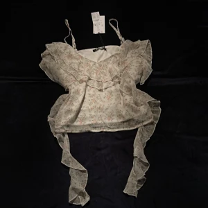 Blommig volangblus från Gina Tricot - Supersöt blommig blus från Gina Tricot i ljus färg med volangdetaljer längs axlar och sidor. Topp med smala axelband, v-ringning och långa volanger som hänger ner. Aldrig använd, prislappen sitter kvar. Säljer pga kommer inte till användning. Den kosta 329kr först, säljer för 230kr.😊❣️