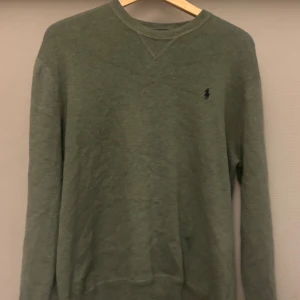 Polo Ralph Lauren - Vintage olivgrön stickad tröja från Polo Ralph Lauren med klassisk rund hals och broderad logga på bröstet. Tröjan är i bomull och har långa ärmar samt ribbade muddar vid hals, ärmslut och nederkant. Perfekt för en chill och stilren look.