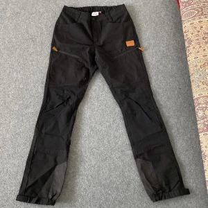 Svarta friluftsbyxor BLWR Woman strl 40 - Svarta friluftsbyxor från BLWR Woman i storlek 40. Byxorna har elastisk midja, förstärkta partier vid knän och benslut, praktiska fickor med dragkedja och justerbara benslut med kardborre. Perfekta för utomhusaktiviteter och äventyr.