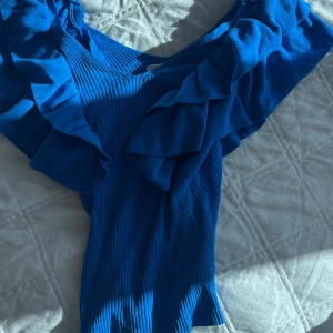 Blå ribbad topp med volang från ONLY - Säljer en blå ribbad topp från ONLY med bred volang längs axlarna och v-ringad hals. Toppen är figurnära och har en snygg struktur som ger extra detalj. Perfekt för dig som vill sticka ut med färg och volym.