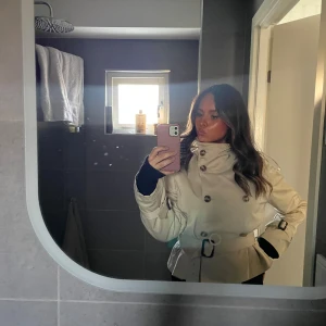 Beige trenchcoat med bälte - Säljer den trendiga beigea korta trenchcoaten från hm med dubbelknäppning, höga axelklaffar och bälte i midjan. Jackan har snygga spännen vid ärmsluten och en cool krage som kan stängas upp. 