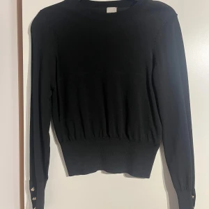 Svart stickad tröja med gulddetaljer H&M - Svart stickad tröja från H&M med lång ärm och bred ribbad midja. guldfärgade knappar på ärmarna. 