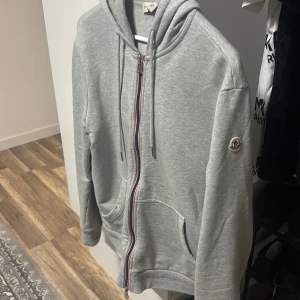 Grå Moncler kofta med dragkedja - Snygg grå hoodie från Moncler med huva och dragsko, hel dragkedja med rödblå detalj och klassisk logga på ärmen. Mjuk bomullskänsla och två fickor framtill.