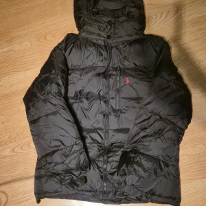 Svart pufferjacka från Polo Ralph Lauren - Svart pufferjacka från Polo Ralph Lauren med röd broderad logga på bröstet. Jackan har huva med snörning, dragkedja framtill och flera fickor med dragkedja. Perfekt för kalla dagar och riktigt snygg streetstil.