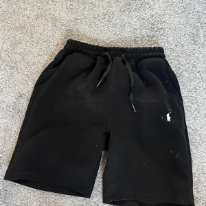 Svarta shorts från Polo Ralph Lauren - Svarta shorts från Polo Ralph Lauren med klassisk vit logga broderad på ena benet. Mjuk bomullskänsla, elastisk midja med snörning och två sidofickor. Perfekta för chill dagar eller när du vill ha en clean och enkel stil.