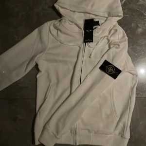 Vit hoodie från Stone Island  - Vit hoodie från Stone Island med klassisk logotyp-patch på ärmen. Tröjan har huva med snörning och hel dragkedja framtill. Tillverkad i mjuk bomull som känns skön mot huden. Perfekt för dig som gillar clean och stilren. Köpte den på Plick för 1500 aldrig använd vet ej om den e äkta skriv vid frågor. Kan sänka pris vid snabb affär 