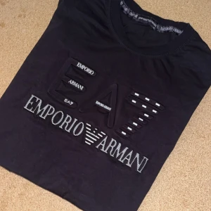 Svart EA7 Emporio Armani t-shirt - Svart t-shirt från Emporio Armani EA7 med stort, upphöjt logotryck och detaljer i vitt på bröstet. Klassisk rund halsringning och korta ärmar. Tillverkad i mjuk bomull för en skön känsla och snygg passform.
