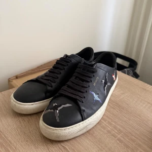 Svarta sneakers från Axel Arigato - Snygga svarta sneakers från Axel Arigato med broderade fåglar i olika färger på sidorna. Skorna har rund tå, svart snörning och vit platt sula. Tillverkade i skinn för en lyxig känsla och cool look. Perfekta för dig som gillar streetstyle och detaljer. Dessa säljs inte längre. Lite smutsiga bara en liten rengöring så är dom dunder! 