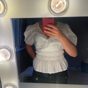 Vit omlottblus från Veromoda - Supersöt vit blus från veromoda med omlottdesign och puffiga korta ärmar. Blusen har smockad midja och volang nedtill som ger en snygg siluett. V-ringad och gjord i mjuk bomull, perfekt för vår och sommar. Passar dig som gillar roma blusar!