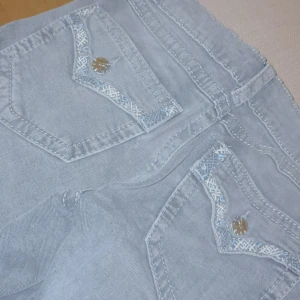 Grå jeansbyxor från Gina Tricot - Snygga grå jeansbyxor från Gina Tricot med dekorativa silvriga knappar och broderade detaljer på bakfickorna. Byxorna har klassisk femficksmodell och raka ben. Perfekta för dig som vill ha en stilren men ändå unik look.