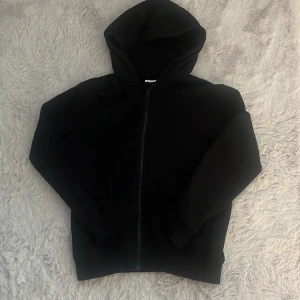Svart hoodie med dragkedja från Kappahl - Svart hoodie från Kappahl i storlek 146/152. Klassisk modell med huva, hel dragkedja framtill och långa ärmar. Perfekt för chill dagar eller när du vill ha en enkel och stilren look.