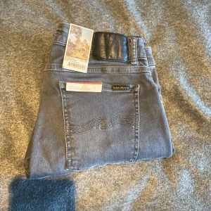Mörkgrå jeans från Nudie Jeans, 32/30 - Snygga mörkgrå jeans från Nudie Jeans med klassisk femficksdesign och diskret broderi på bakfickorna. Modellen har en smal passform och är tillverkad i mjukt jeanstyg med stretch för extra komfort. Perfekta för dig som gillar stilrena och moderna jeans.
