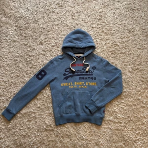 Blå hoodie från superdry - Snygg blå hoodie från Superdry med snygga tryck, både på bröstet och ärmen. Den är i mycket gott skick förutom att trycket har spruckit lite vilket syns på bilderna om man kollar noga, men det är inget som gör den mindre snygg enligt mig. Storlek S, tveka inte på o skriva vid frågor!
