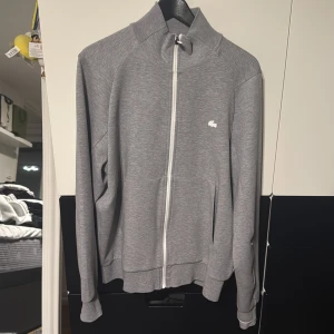 Grå ziptröja från Lacoste - Snyggt grå set från Lacoste i klassisk passform. Tröjan har hel dragkedja framtill, ståkrage och två fickor. Liten broderad Lacoste-logga på bröstet. Perfekt för lager-på-lager och chill vardagsstil medans byxan är i klassisk grå färg som matchar tröjan. Använda 1-2 ggr nypris 2800