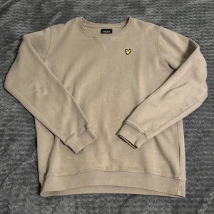 Beige sweatshirt från Lyle & Scott - Snygg beige sweatshirt från Lyle & Scott med klassisk rund hals och gul logga på bröstet. Tröjan har ribbade muddar vid ärmslut och nederkant samt lång ärm. Perfekt för en clean och stilren look.