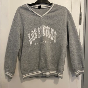 Grå college-tröja Los Angeles  - Grå college-tröja från i storlek XS med vit text 'Los Angeles California' framtill. V-ringad med vita och grå ränder vid hals, ärmslut och nederkant. Mjuk insida och långärmad. Passar både Xs och S. knappt använd.