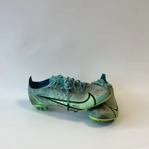 Nike Mercurial fotbollsskor turkos - Nike Mercurial fotbollsskor i turkos och limegrön med svart swoosh. Skorna har snörning, platt sula med dobbar och är tillverkade i syntetmaterial med meshstruktur. Snygg sportig design med rund tå och detaljerade mönster på ovansidan. Perfekta för dig som älskar fotboll.