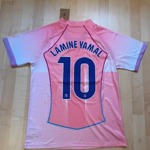 Barça Lamine Yamal rosa matchtröja - Säljer en officiell FC Barcelona fotbollströja från Nike med Lamine Yamal och nummer 10 på ryggen. Tröjan är rosa med vita och lila detaljer, har korta ärmar och ett grafiskt mönster framtill. Materialet är lätt polyester, perfekt för träning eller match.