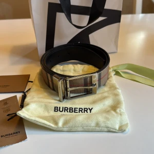 Burberry vändbart bälte - Säljer ett vändbart Burberry-bälte med klassiskt rutigt mönster i beige, svart och rött på ena sidan och helsvart skinn på andra. Snyggt spänne i silverfärgad metall med Burberry-logga. Perfekt accessoar för att lyfta din outfit. Pris inte hugget i sten, bara att skriva om de är något du undrar över.