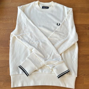 Vit/beige sweatshirt från Fred Perry M - Snygg vit/beige sweatshirt från Fred Perry i bomull med svart broderad logga på bröstet. Tröjan har rund hals, ribbade muddar med svarta ränder och klassisk passform. Perfekt för en clean och stilren look.