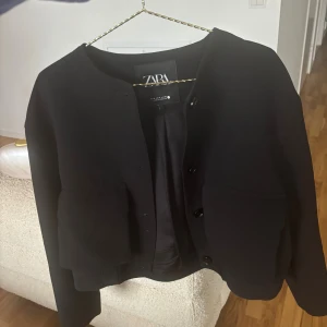 Svart cardigan jacka från Zara - Snygg svart cardigan jacka från Zara med rund halsringning och stora fickor framtill. Jackan har knappar och är långärmad med ribbade muddar. Perfekt för dig som gillar stilrena och enkla plagg med en modern touch.