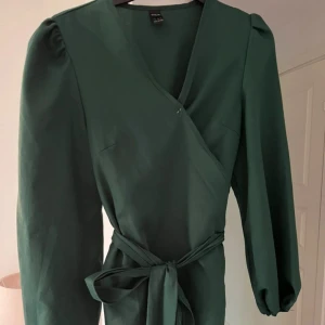 Mörkgrön omlottklänning med bälte - Supersnygg mörkgrön omlottklänning från SHEIN med långa puffärmar och knytband i midjan. Klänningen har v-ringad hals och en stilren, elegant look som passar dig som gillar att sticka ut med färg. Materialet är mjukt och känns skönt mot huden.