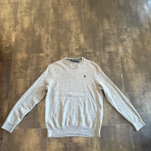 Ralph Lauren tröja - Snygg ljusgrå tröja från Ralph Lauren med rund hals och diskret broderad logga på bröstet. Tröjan är stickad i mjukt material och har långa ärmar samt ribbade muddar vid ärmslut och nederkant. Perfekt för en clean och klassisk stil.