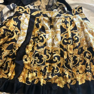 Versace långkjol med barockmönster - Säljer en maxad långkjol från Versace med ikoniskt barockinspirerat mönster i guld, svart och vitt. Kjolen har breda veck, dragkedja och knappar i midjan. Materialet är mjuk polyester som ger ett lyxigt fall. Perfekt för dig som vill sticka ut med en statement piece.