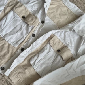 Beige och vit jeansjacka med fickor - Snygg jeansjacka i beige och vitt med kontrasterande paneler. Jackan har två stora bröstfickor med lock och knappar, klassisk krage och knäppning framtill. Perfekt för dig som gillar en clean och stilren look med lite edge.