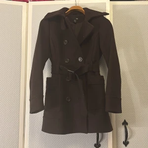 Brun kappa från H&M med bälte - Snygg brun trenchcoat från H&M i klassisk modell med dubbelknäppning, stora fickor och bälte i midjan. Jackan har bred krage och långa ärmar, perfekt för höst och vinter. Materialet är ullblandning som håller dig varm och stilen är tidlös.