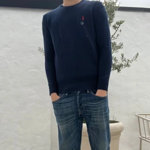 Mörkblå stickad tröja (Helt ny) - (Modellen är 183)Snygg mörkblå stickad tröja från Polo Ralph Lauren med klassisk rund hals och röd broderad logga på bröstet. Tröjan har ribbade muddar vid ärmslut och nederkant, och är långärmad. Perfekt för dig som gillar stilrena och tidlösa plagg.