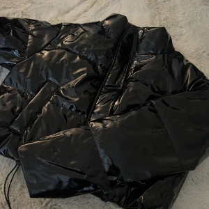 Svart glansig pufferjacka med dragkedja - Svart pufferjacka med glansig finish. Jackan har dragkedja och tryckknappar framtill samt fickor på sidorna. Inte den varmaste jackan, detta är mer en höst jacka.
