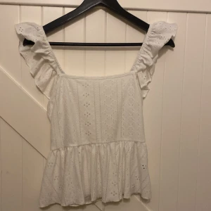 Vit blus med volang och broderi - Superfin vit blus med breda axelband och volangdetaljer. Blusen har broderade hålmönster och en söt knytning i ryggen. Den är lätt och luftig med en öppen rygg, perfekt för varma dagar. 