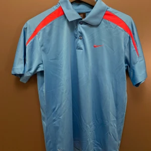 Blå Nike pikétröja med röda detaljer - Säljer en blå pikétröja från Nike med röda detaljer på axlarna och liten röd swoosh-logga på bröstet. Tröjan har klassisk krage, knappar framtill och är tillverkad i lätt polyester med Nike FITDRY-teknologi. Perfekt för sport eller chill dagar.