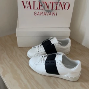 Valentino Garavani vita sneakers - Snygga vita sneakers från Valentino Garavani med bred svart detalj över snörningen. Skorna har rund tå, platt sula och är tillverkade i skinn. Perfekta för dig som gillar stilrena och exklusiva sneakers med en unik touch.