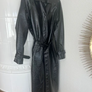 Svart lång kappa i svart lackliknande material från NLY Trend - Snygg svart mycket trendig lång trenchcoat  i skinnimitation i lack liknande material från NLY Trend. Kappan har långa ärmar, knappar framtill och bälte i midjan för en markerad siluett. Perfekt för dig som vill sticka ut med en edgy look.