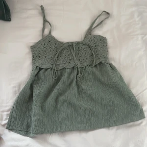 Grön linnetopp från Stradivarius XS - Supersöt grön linnetopp från Stradivarius i storlek XS. Toppen har virkade detaljer upptill, smala axelband och knytning framtill. Nederdelen är i ett mjukt, lätt skrynklat tyg som ger en chill vibe. Perfekt för varma dagar och sommarhäng.